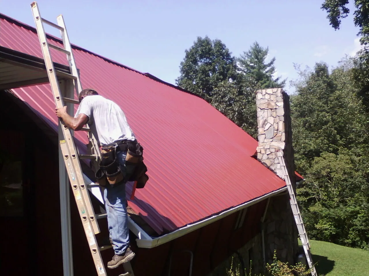 Roofer on a ladder beside a red metal roof and stone chimney — Soffit & Fascia Repair by Roofing Brothers in Ringtown